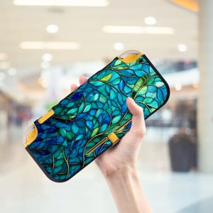 Könnte beinhalten: Eine Hülle für ein tragbares Spielgerät mit einem blau-grünen Blättermuster. Die Hülle hat goldene Details und gelbe Bedienelemente. Das Design zeigt ein Mosaik aus blauen und grünen Blättern mit goldenen Umrissen.