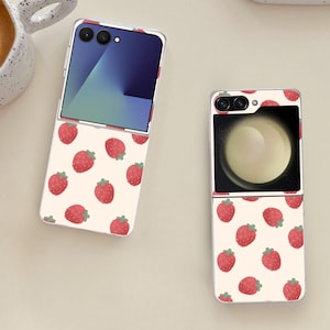 May include: Two open and closed foldable smartphones with a strawberry pattern on a cream background. The strawberries are red with green stems. The phone screens display a blue and white gradient and a light-colored circle.
