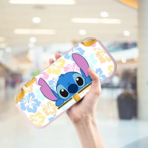 May include: A white and pink Switch Lite case featuring a blue cartoon character with large ears and eyes, surrounded by a floral pattern in yellow, pink, and blue. The case has yellow accents.