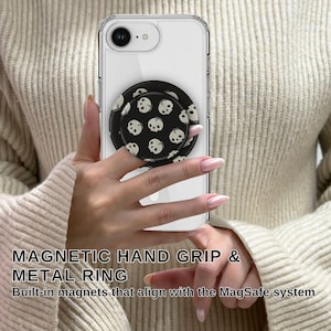 May include: A clear phone case with a black and white skull design phone grip and metal ring. The phone grip is attached to a white phone. The text on the image reads "MAGNETIC HAND GRIP & METAL RING".