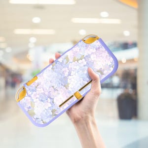 Puede incluir: Una funda para consola de juegos portátil con un estampado floral en lavanda y blanco. La funda tiene detalles en amarillo y un borde lavanda. El diseño floral presenta flores blancas y lavanda con hojas verdes.