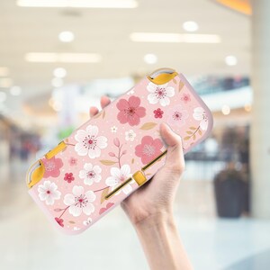 May include: A pink and white floral patterned Nintendo Switch Lite case. The case features a repeating pattern of pink and white flowers with yellow accents. The case is designed to protect the Switch Lite console.