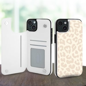 Puede incluir: Dos fundas para teléfono. Una es blanca con un diseño de cartera, con ranuras para tarjetas y un cierre magnético. La otra funda tiene un diseño de estampado de leopardo beige. Ambas fundas tienen un borde negro y están sobre una superficie de madera.