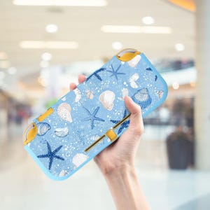 May include: A light blue handheld gaming console case featuring a pattern of white seashells and blue starfish. The case has yellow accents and a light blue border. The design is reminiscent of a beach theme.