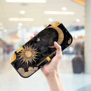 Può includere: Una custodia per Switch nera e dorata con un design celeste. La custodia presenta un sole, una luna, stelle e nuvole. La custodia ha dettagli gialli ed è tenuta in mano.