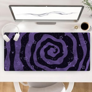 May include: A rectangular desk mat featuring a dark purple and black swirling spiral pattern with scattered white star outlines and small white dots. The mat is positioned on a white desk surface below a computer monitor and white headphones.