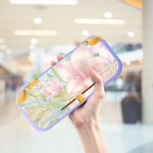 Può includere: Una custodia per Switch Lite lilla e oro con un design floreale. Presenta fiori rosa e bianchi, foglie verdi e dettagli dorati. Ha un bordo protettivo e pulsanti dorati.