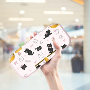 Könnte beinhalten: Eine hellrosa und weiße Nintendo Switch-Hülle mit einem sich wiederholenden Muster aus schwarzen Katzen, Schleifen, Eiswaffeln und anderen niedlichen Illustrationen. Die Hülle hat gelbe Akzente und einen rosa Rand.