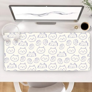 Peut inclure: Tapis de bureau rectangulaire crème avec un motif répétitif de visages de chats bleus et d'empreintes de pattes. Le tapis est posé sur un bureau blanc, avec un écran d'ordinateur, un casque et une tasse de café visibles.