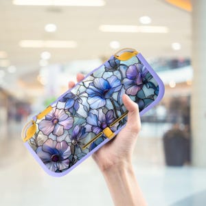 Könnte beinhalten: Eine lavendelfarbene Nintendo Switch Lite Hülle mit einem Blumendesign in Blau- und Lilatönen. Die Hülle hat gelbe Akzente und eine glänzende Oberfläche. Das Blumenmuster erinnert an Buntglas.