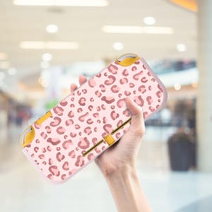 May include: A pink and brown leopard print Nintendo Switch Lite case. The case has a light pink background with brown leopard spots. The buttons and accents are yellow. The case is held in a person's hand.