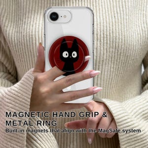 May include: A clear phone case with a black cat design on a red circular magnetic hand grip. The text "MAGNETIC HAND GRIP & METAL RING" is visible. The phone case is held in a person's hands.