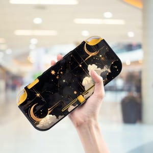 以下が含まれることがあります： 黒と金のNintendo Switch Liteケース。星、月、雲のような形が描かれた黒い背景に、金色のデザインが施されています。ケースの側面は黄色です。
