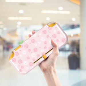 Può includere: Una custodia rosa per console di gioco portatile con un motivo floreale bianco e rosa. La custodia presenta bottoni e dettagli dorati. Lo sfondo è sfocato, suggerendo un ambiente interno.