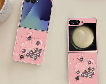 以下が含まれることがあります： 黒いススワタリで満たされたハート型のデザインのピンク色のフリップ携帯電話ケース。ケースは開いており、電話の画面が表示されています。デザインには小さな白い点と追加のススワタリのイラストが含まれています。