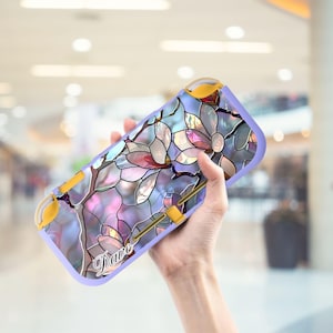 Könnte beinhalten: Eine lavendelfarbene Spielkonsolenhülle mit einem Buntglas-Blumendesign in Rosa, Weiß und Blau. Die Hülle hat gelbe Akzente und der Name "Dave" steht unten. Die Hülle wird von einer Person in der Hand gehalten.
