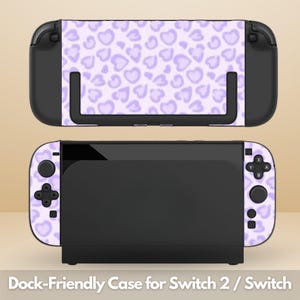 Pode incluir: Uma capa para console de jogos com um padrão de corações em violeta claro. O dispositivo é exibido em duas vistas: uma encaixada em uma estação de carregamento preta e outra separada, mostrando os botões e joysticks pretos sobre a estampa.