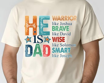 Op de afbeelding: Crèmekleurig T-shirt met de tekst "HE IS DAD" in kleurrijke, verweerde letters. Het shirt vermeldt ook eigenschappen als "WARRIOR" en "BRAVE". Een casual, comfortabel T-shirt voor Vaderdag of elke dag.