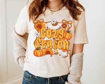 May include: Cream-colored t-shirt with the words "Cozy Season" in orange bubble letters. The design includes pumpkins, sunflowers, and daisies. The shirt is a casual, fall-themed apparel item.