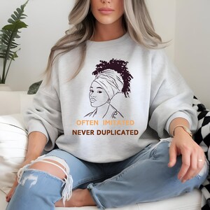 May include: A gray sweatshirt with a black and white line drawing of a woman with a headscarf. The text "Often Imitated Never Duplicated" is printed below the drawing.