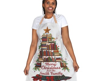 Op de afbeelding: Witte schort met een kerstboomontwerp gemaakt van boeken, een ster en feestelijke lichtjes. De schort bevat de tekst "Merry Christmas from the Smith Family" en een ruitpatroon aan de onderkant met een rode strik.