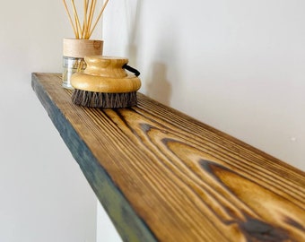 May include: A dark brown wooden shelf with a natural wood grain finish. The shelf has a black edge and is mounted on a white wall. A glass jar with wooden lid and wooden sticks is on the shelf, along with a wooden brush.