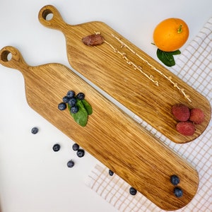 May include: Two wooden chopping boards with handles. The boards are made of light brown wood and have a natural finish. One board has a few blueberries and a sprig of green leaves on it. The other board has a few dried dates and a few dried flower buds on it. Both boards are on a white surface with a white and brown checkered cloth.