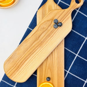 May include: Two wooden serving boards with handles. The boards are made of light wood and have a natural finish. One board has three blueberries on it, and the other has a slice of orange and some green leaves.