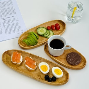 May include: Three wooden serving trays with various snacks and drinks. The trays are oval shaped and made of light brown wood. The snacks include avocado slices, cucumber slices, cherry tomatoes, salmon on bread, hard-boiled eggs, and a chocolate cookie. The drinks include a cup of coffee and a glass of water with a lemon slice.