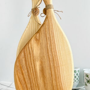 May include: Two wooden chopping boards with handles, one on top of the other, with a small wooden stand. The boards are light brown and have a natural wood grain. The handles are shaped like a teardrop and have a hole in the top. The boards are tied together with twine and a small tag that says "Handmade with love".