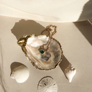 May include: A gold-rimmed oyster shell dish with a gold chain necklace and a green gemstone pendant. The dish is sitting on a beige surface with a white seashell and a sand dollar.