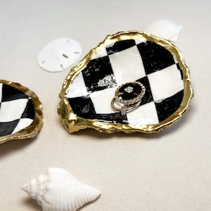 May include: A black and white checkered oyster shell ring dish with a gold rim. The dish is filled with two gold rings.