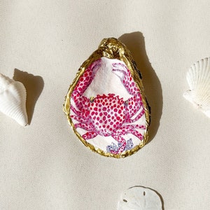 May include: A gold-painted oyster shell with a pink and white crab design. The crab has red polka dots and is surrounded by blue and purple seaweed.
