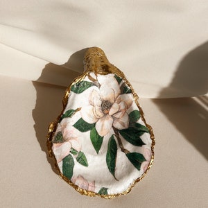 May include: A white oyster shell with a gold rim, decorated with a floral pattern of pink and white magnolia flowers and green leaves.