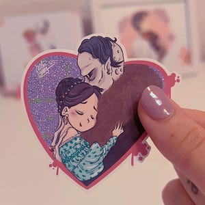 Op de afbeelding: Een sticker met een hartvormig ontwerp en een paarse bloemenachtergrond. In het hart is een cartoonkoppel afgebeeld in een liefdevolle omhelzing. Het mannelijke personage heeft blauw haar en een paarse, harige jas. Het vrouwelijke personage heeft lang, donker haar en draagt een blauwe en witte jurk.