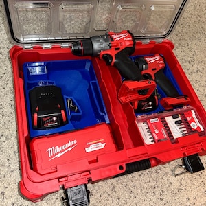 May include: A red Milwaukee tool case with a blue insert. The case contains two red and black cordless drills, a black battery, and a clear plastic container of drill bits.