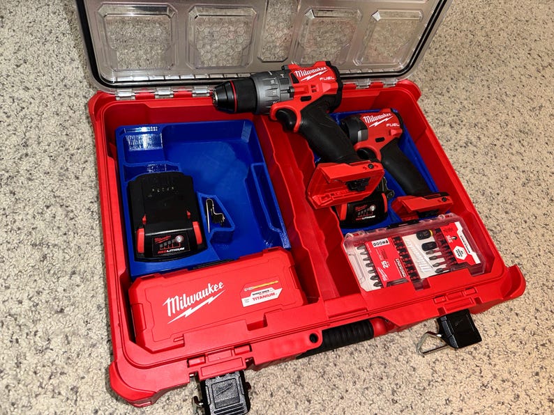 May include: A red Milwaukee tool case with a blue insert. The case contains two red and black cordless drills, a black battery, and a clear plastic container of drill bits.