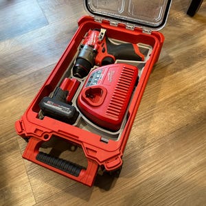 May include: A red power tool kit with a clear lid, containing a drill, a battery, and a charger. The kit is open, revealing the tools neatly arranged inside. The handle is black.