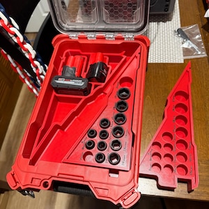 May include: A red toolbox with a clear lid, open to reveal various tools. Inside are sockets, a battery, and other components. The toolbox is on a wooden surface.