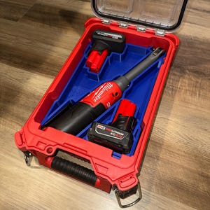 May include: A red Milwaukee tool case with a clear lid, containing a red and black power tool, two batteries, and a charger. The interior is blue. The case is open, revealing the tools inside.