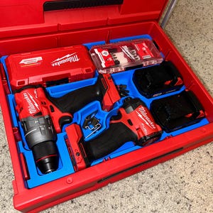 May include: A red toolbox with a blue foam insert. The toolbox contains two Milwaukee power drills, two batteries, and a set of drill bits. The toolbox is open and the contents are visible.