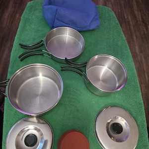 May include: A stainless steel camping cookware set with black handles and lids. The set includes several pots and pans, a red lid, and a blue carrying bag. The cookware is arranged on a green towel.