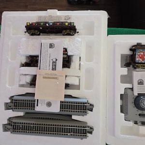 May include: A new, boxed model train set featuring several train cars, track pieces, and a power supply. The train cars have festive holiday decorations. The set is packaged in a white styrofoam container.