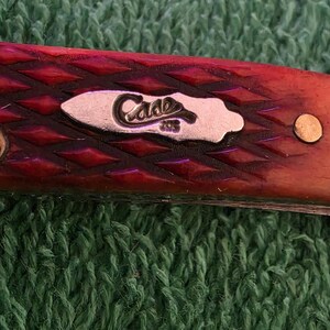 May include: Close-up of a red pocket knife with a diamond pattern on the handle. The knife features a silver emblem with the word "Case" and the number "208". The knife is set against a green textured background.