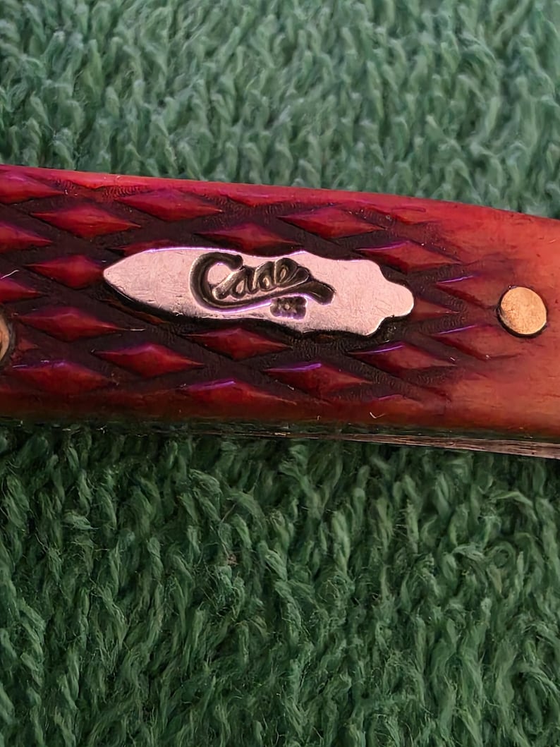 May include: Close-up of a red pocket knife with a diamond pattern on the handle. The knife features a silver emblem with the word "Case" and the number "208". The knife is set against a green textured background.