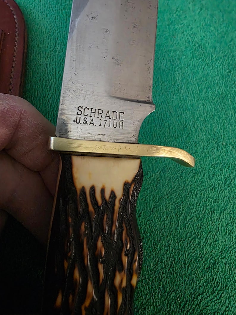 Pu&ograve; includere: Un coltello da caccia della marca Schrade con un manico in corno di cervo marrone e nero e una guardia in ottone. La lama &egrave; stampata con "Schrade U.S.A. 171UH".