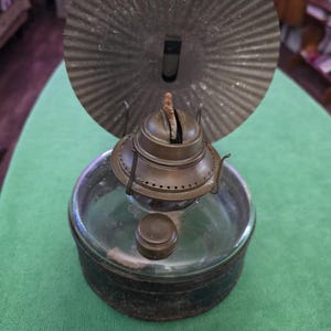 May include: A vintage oil lamp featuring a clear glass base and a dark metal burner assembly. A large, circular, corrugated metal reflector is attached to the back. The lamp includes a small, round metal filler cap and a visible wick at the top.