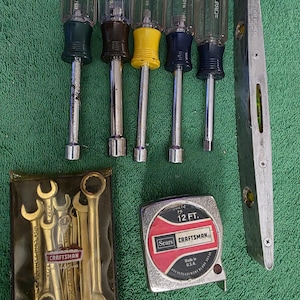 May include: A collection of Craftsman tools, including nut drivers with colored handles, a level, a tape measure, and a set of wrenches. The tape measure reads 12 FT. and the tools are on a green surface.