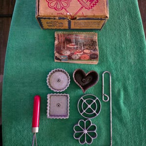 May include: Vintage Double Rosette and Timbal Iron set. The set includes a box with illustrations of rosette and heart-shaped cookies, along with various metal cookie molds and a red-handled tool. The box reads "DOUBLE ROSETTE and TIMBALE IRON".