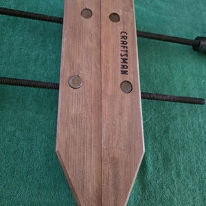 May include: A Craftsman wood clamp with a dark wood finish. The clamp has two wooden jaws, metal hardware, and a threaded rod for adjusting pressure. The Craftsman logo is printed on the side.